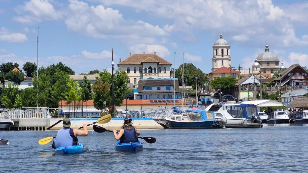Doi caiaciști vâslind pe lacul Razim, cu portul, bărcile și turlele bisericii din Jurilovca în fundal
