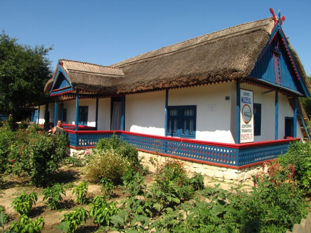 Casa albă a Gospodăriei Țărănești din Enisala, cu acoperiș gros de stuf și pridvor cu balustradă albastră și roșie, văzută din grădina de legume din față