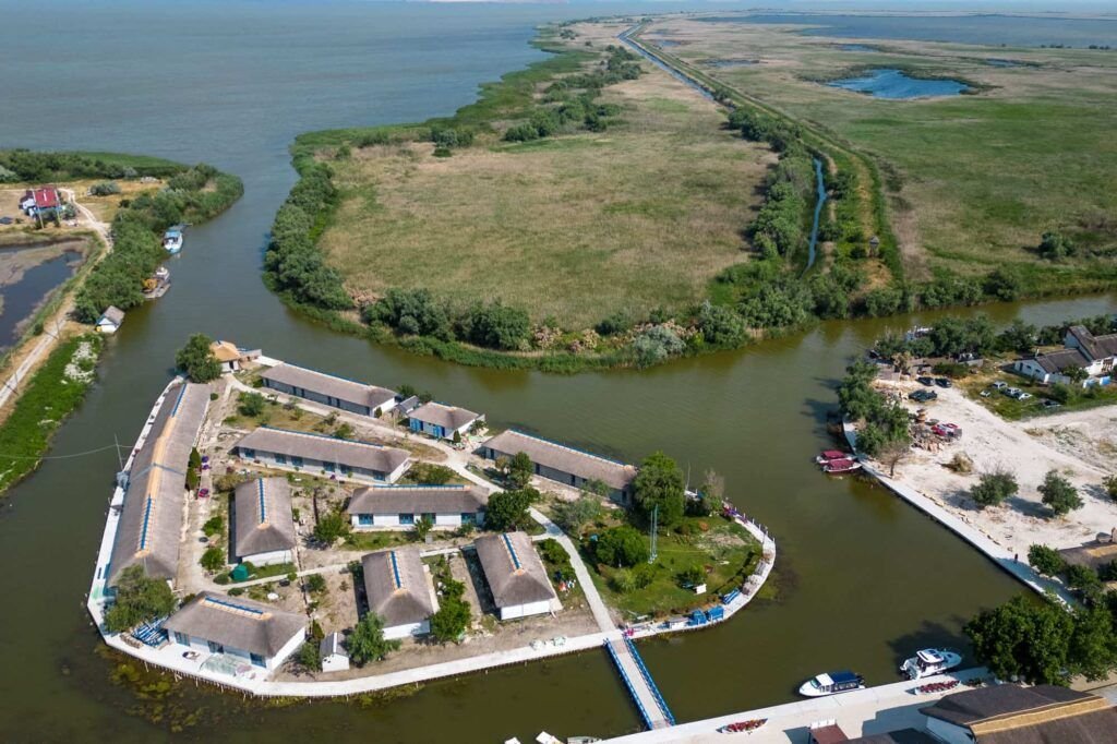 Vedere de sus asupra căsuțelor cu acoperiș de stuf de la Gura Portiței, așezate pe fâșia de pământ dintre lagună și Marea Neagră, cu pontonul și bărcile alături