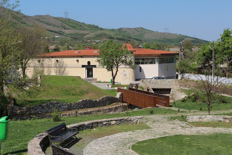 Clădirea-muzeu care adăpostește bazilica paleocreștină de la Niculițel, cu acoperișuri de țiglă roșie, o pasarelă de lemn și dealurile împădurite în spate