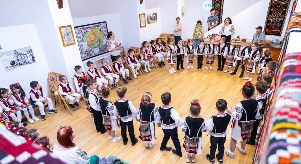 Grup de copii în costume populare așezați și stând în cerc într-o sală a muzeului, cu țesături și covoare tradiționale pe pereți