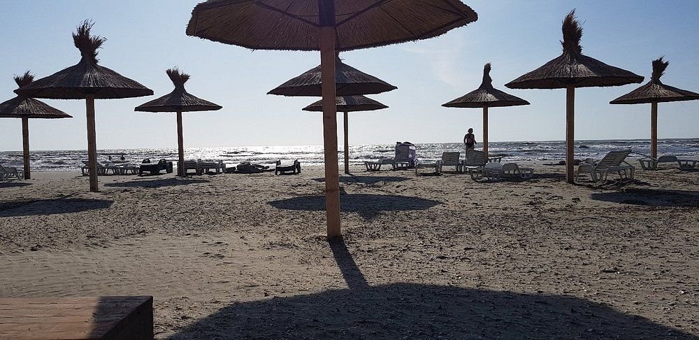 Umbrele de stuf și șezlonguri pe nisipul plajei de la Sulina, cu marea în contralumină și cățiva oameni la mal