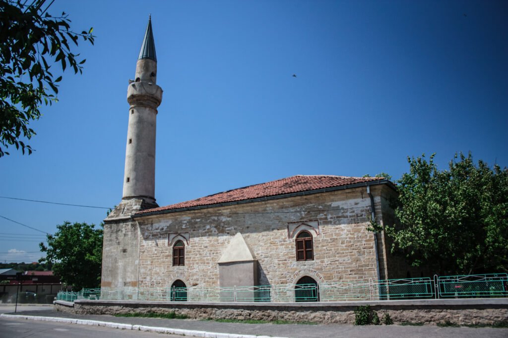 Geamia Ali Gazi Pașa din lateral: ziduri de piatră, acoperiș de țiglă roșie și minaretul de piatră cu vârf ascuțit, pe cer albastru