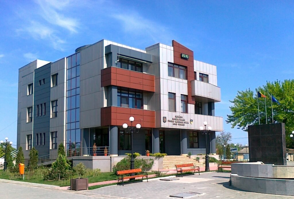 Primăria din Isaccea: clădire modernă cu fațadă gri și roșu-cărămiziu, cu steaguri, felinare, bănci și o fântână în piațeta din față, cer albastru