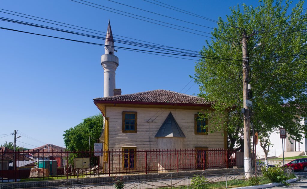 Geamia Mestan Aga din Măcin: clădire cu pereți crem, ferestre cu rame galbene, acoperiș de țiglă și minaret alb, după un gard roșu
